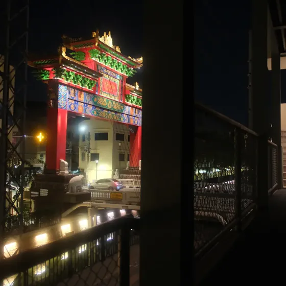 An illuminated Chinatown gate at night, seen through a window past a dark railing